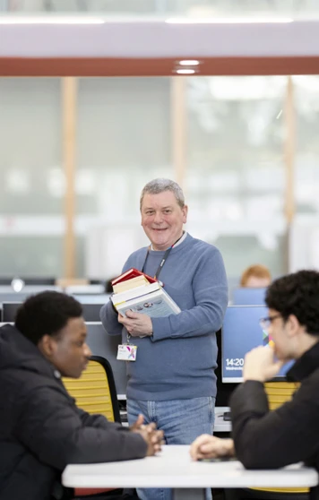 Smiling staff member holding books and standing beside two students in a Glasgow Kelvin College library study pod. Smiling staff member holding books and standing beside two students in a Glasgow Kelvin College library study pod.